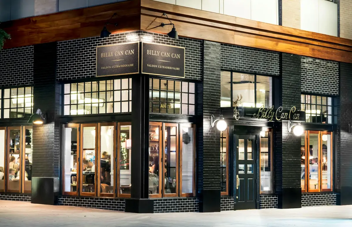 Billy Can Can at night — black brick corner facade with brass Saloon Extraordinaire signage and warmly lit windows